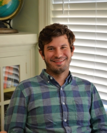 Professor Justin Cook sits in front of a bookshelf and blinds, posing for a photograph.